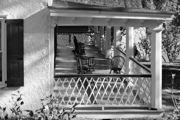 Black-and-white photograph of a porch with wooden chairs.